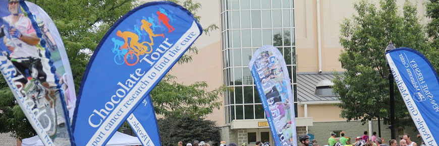 An outdoor banner/sign for the Chocolate Tour featuring silhouettes of cyclists and runners, stands among several others for the tour in an outdoor setting.