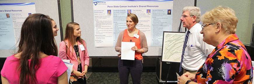 A young female medical student, approximately in her twenties, stands in front of a poster for Penn State Cancer Institute’s Shared Resources, addressing three other young female medical students, also in their twenties, and two adults approximately in their fifties.