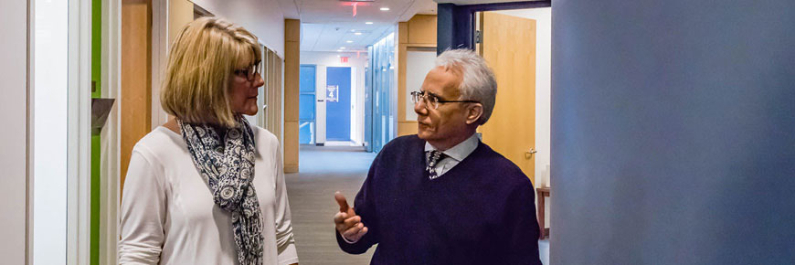 An older male walks and talks with a female in a hospital hallway.