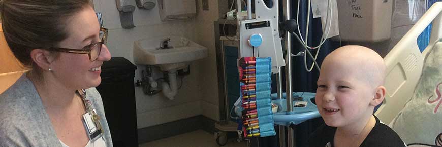 Young child sitting up in their hospital bed speaking with a medical provider, colored crayon hanging off an IV pole.