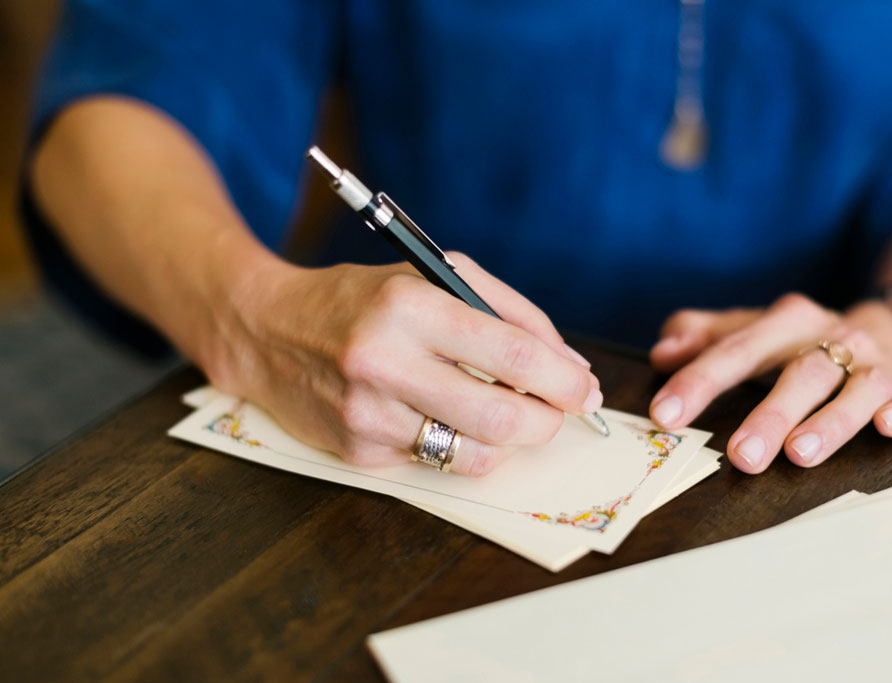 A woman holds a pen and writes on notepaper lying on a table. Several blank sheets of paper sit on the table in front of her.