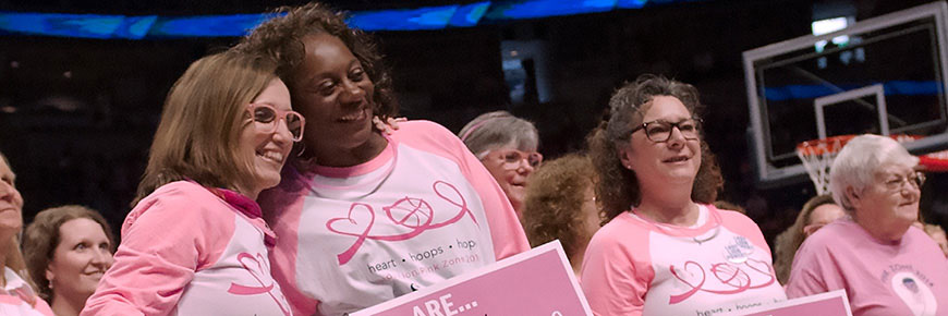 Two women in matching pink shirts pose together for a photo while several other women also in pink tee shirts stand nearby.