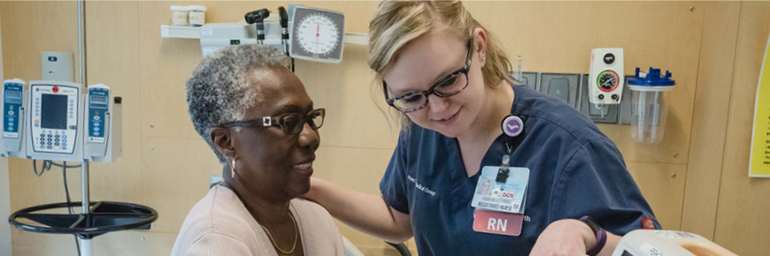 A female RN in blue nurses uniform speaks with an older African American woman in a hospital room.