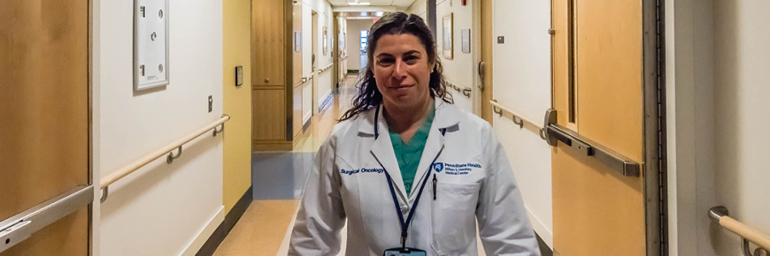 A female doctor in a white coat and scrubs walks in a hospital hallway.