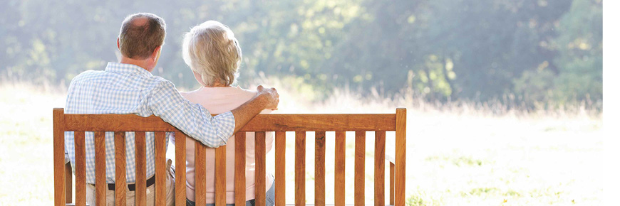 Older man and woman sit on a wooden park bench outdoors looking at the view in front of them of an open field and trees.