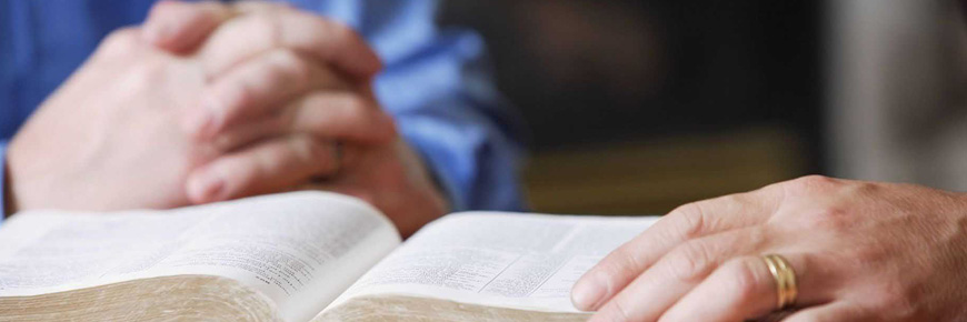 Close up of a hand with a wedding band about the flip a page of the Bible, with another set of hands in the background.