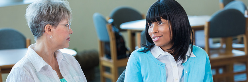 Two women talk together in a cafeteria setting.