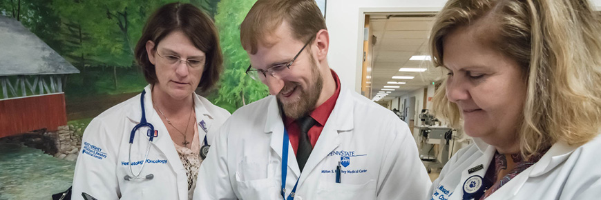 Three doctors, one male and two female, in a hospital hallway, smile and laugh while looking down at something.