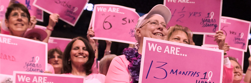 A group of women all wearing pink and holding fill-in-the-blank pink signs that say “We ARE …” stand together.