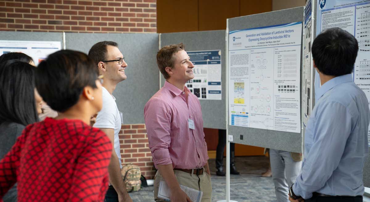 Three men and two woman are pictured talking to each other while gesturing at an academic poster.